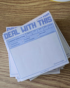 A stack of white notepad sheets with blue printed text and fields, placed on a wooden tabletop next to a cup of coffee.