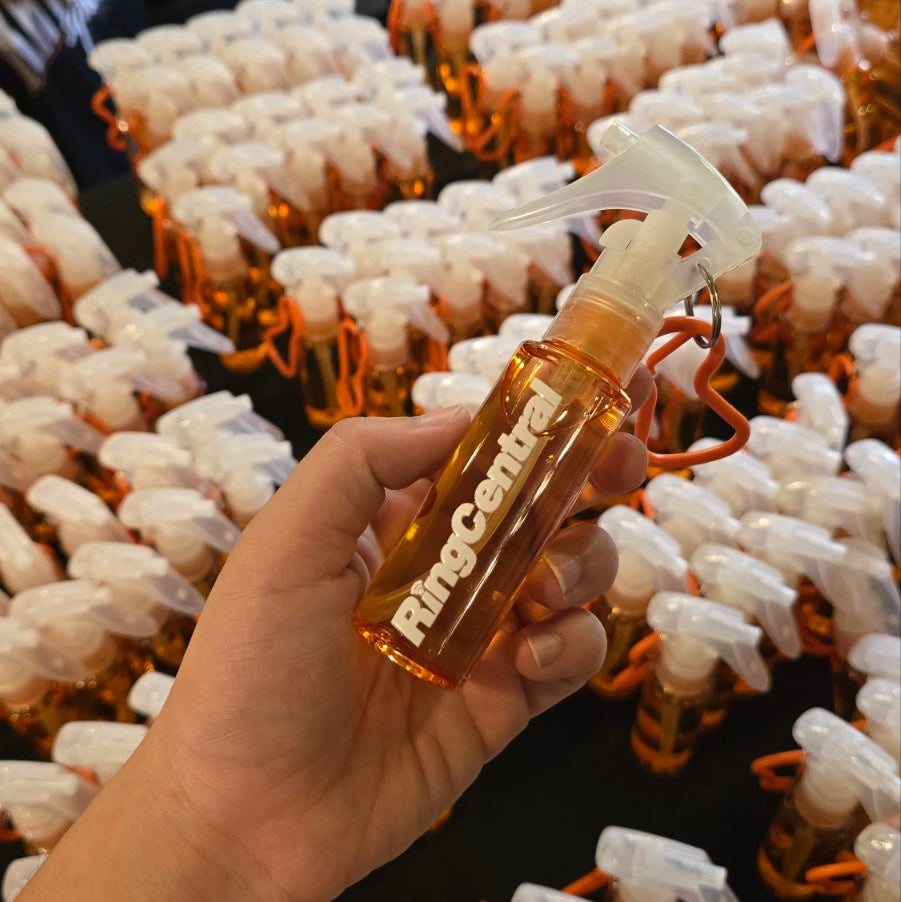 A hand holds a small amber spray bottle with a clear trigger sprayer and orange loop, branded 'RingCenter', in front of a table covered with many identical bottles.