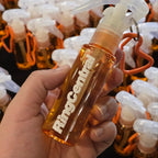 A hand holds a small clear plastic spray bottle filled with amber liquid, labeled RingCentral, with a white trigger sprayer and orange keychain loop. More identical bottles are visible in the blurred background.