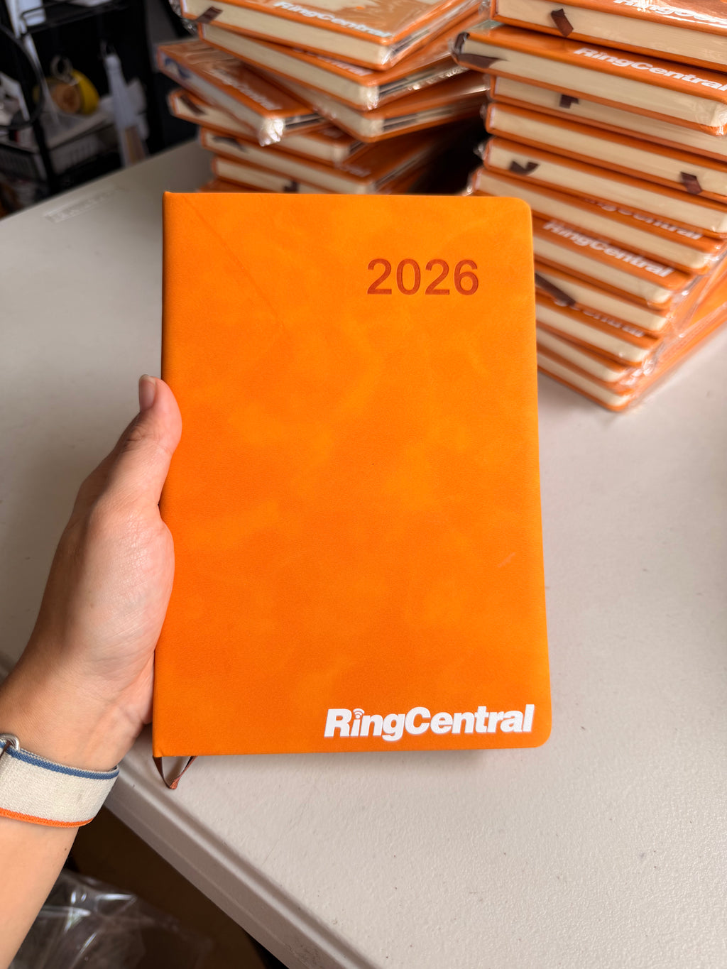An orange hardcover 2026 planner held by a hand, with stacks of similar orange planners in the background on a white table. The brand name 'RingCentral' is printed in white at the bottom of the planner cover.