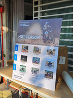 A large promotional poster for BMW Motorrad 'Hot Season Hotter Deals' displayed on a wooden table in a real-world setting with a cardboard box behind it.