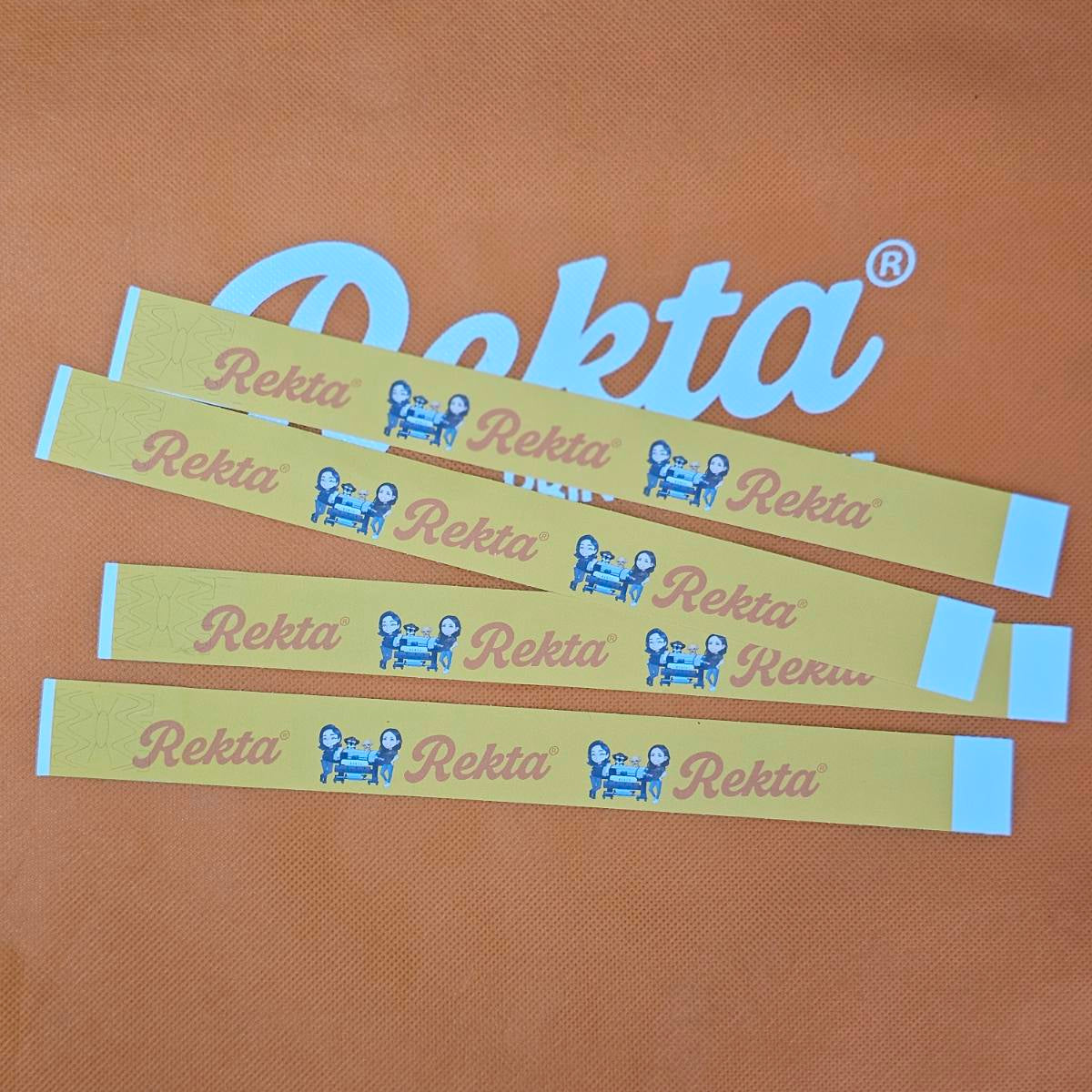 Four yellow paper wristbands with blue and pink Rekta branding are laid out on an orange fabric surface with a large white Rekta logo in the background.
